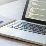 Laptop with code editor open, notebook and pen on wooden desk, perfect for tech and work themes.