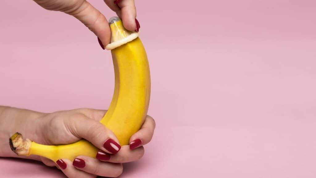 Close-up of hands applying a condom on a banana against a pink background symbolizing safe sex.