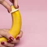 Close-up of hands applying a condom on a banana against a pink background symbolizing safe sex.