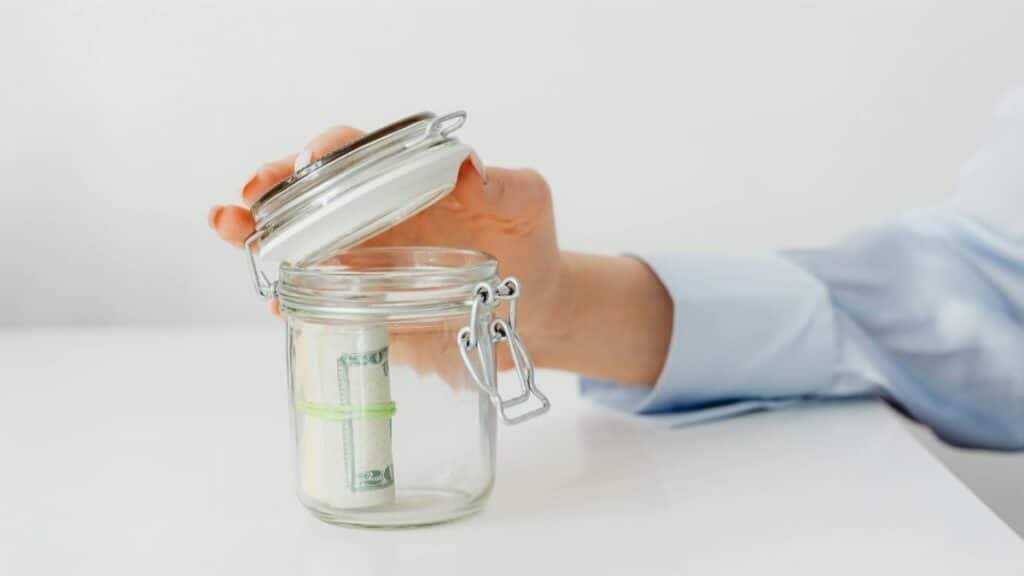A hand places money in a glass jar on a white table, symbolizing savings.