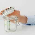 A hand places money in a glass jar on a white table, symbolizing savings.