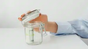 A hand places money in a glass jar on a white table, symbolizing savings.