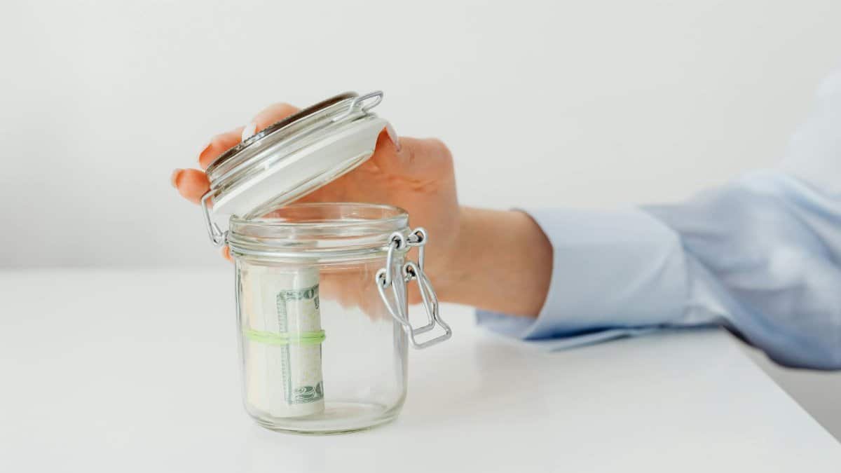 A hand places money in a glass jar on a white table, symbolizing savings.