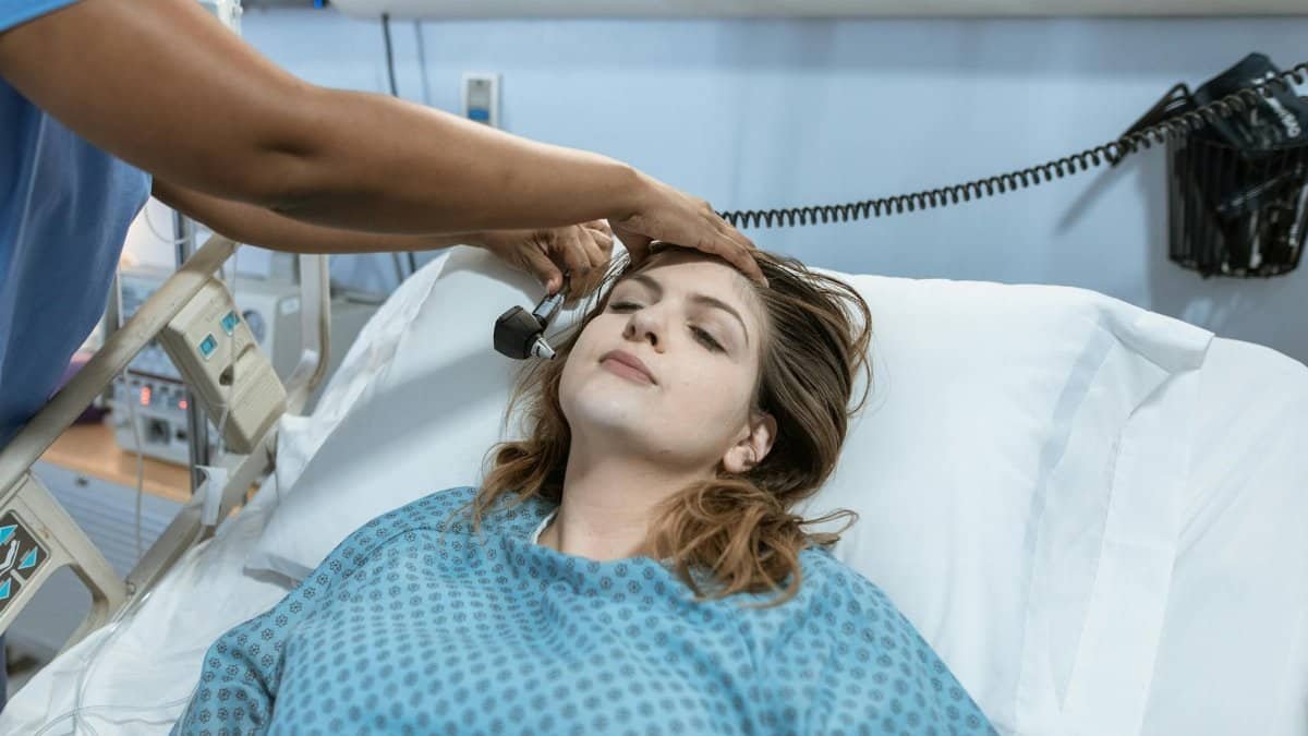A female patient in a hospital bed undergoing a check-up with a healthcare worker using an otoscope.