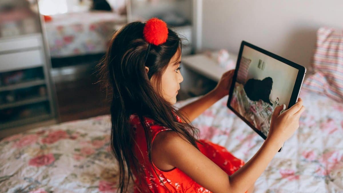 Child in red dress using a tablet in a cozy bedroom. Capturing tech interaction.