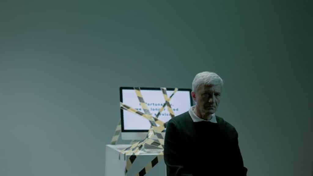 A senior adult sits indoors next to a computer wrapped in caution tape, symbolizing job loss and unemployment.