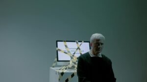 A senior adult sits indoors next to a computer wrapped in caution tape, symbolizing job loss and unemployment.