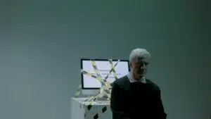 A senior adult sits indoors next to a computer wrapped in caution tape, symbolizing job loss and unemployment.