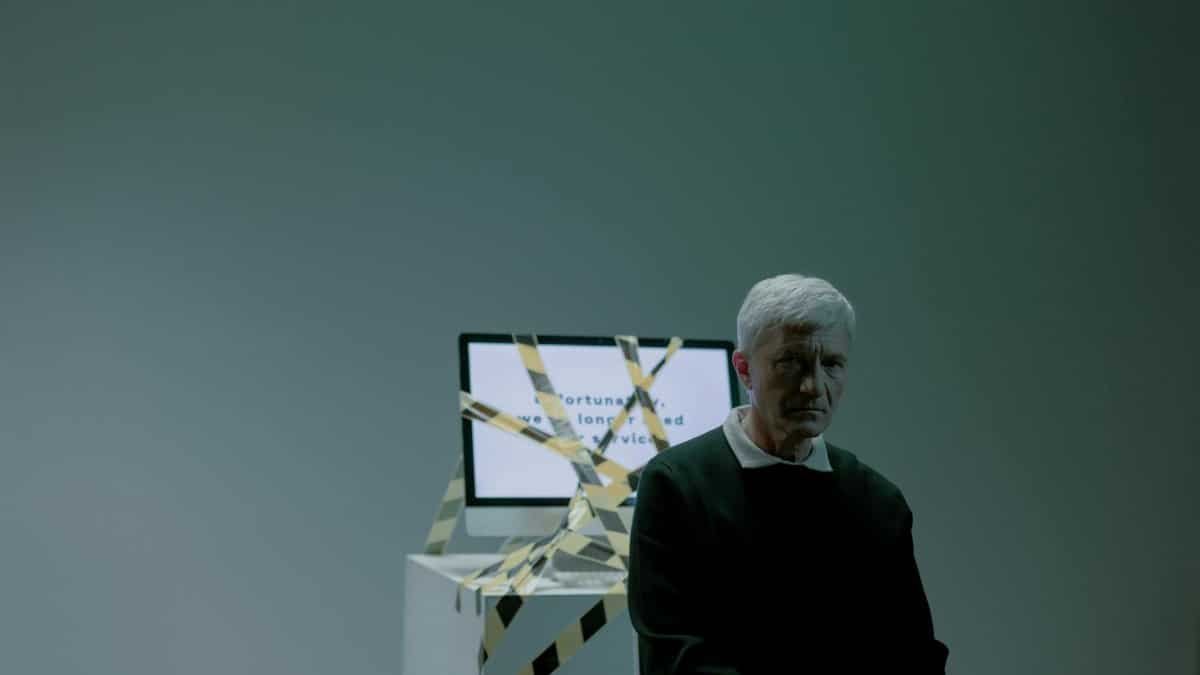 A senior adult sits indoors next to a computer wrapped in caution tape, symbolizing job loss and unemployment.