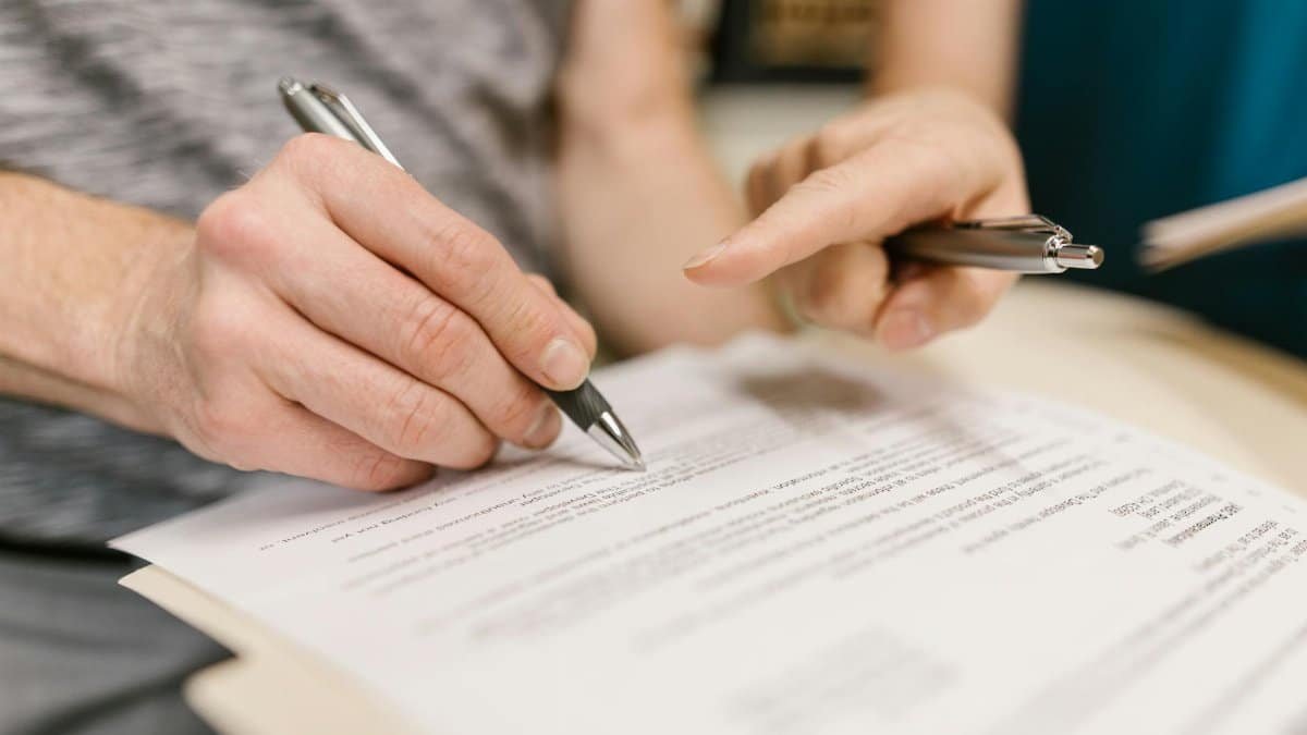 Close-up of two adults reviewing and signing a legal document with pens indoors.