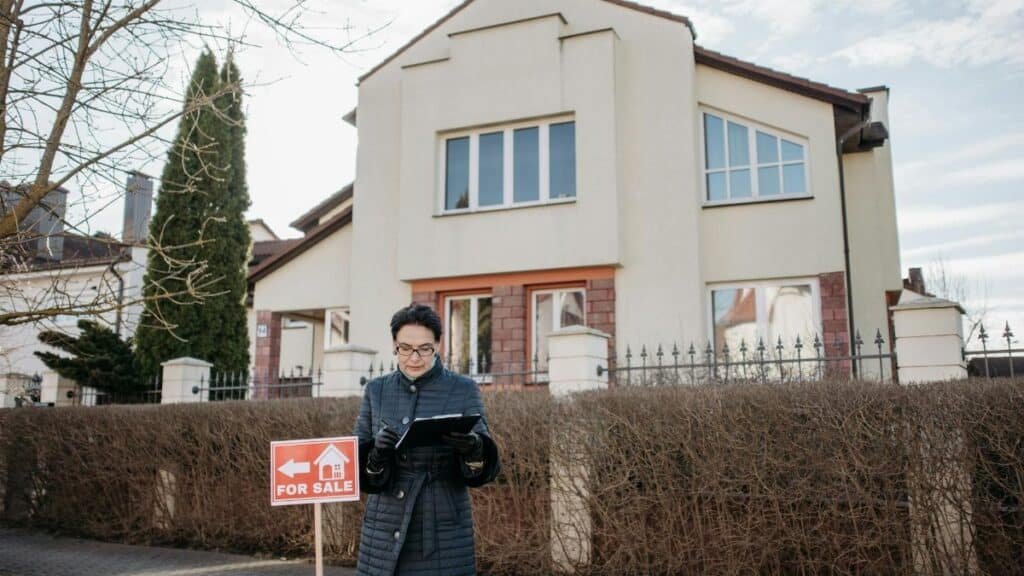 A real estate agent standing outside a house with a 'For Sale' sign, ready for viewing.