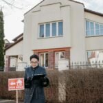 A real estate agent standing outside a house with a 'For Sale' sign, ready for viewing.