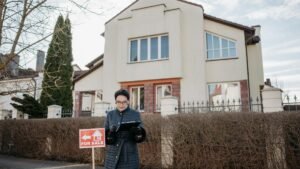 A real estate agent standing outside a house with a 'For Sale' sign, ready for viewing.
