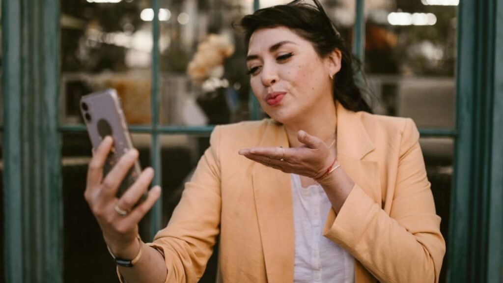 A woman in a beige blazer blows a kiss while talking on a video call outdoors.