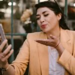 A woman in a beige blazer blows a kiss while talking on a video call outdoors.