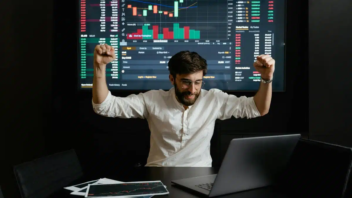 Businessman celebrates stock market success with hands raised in excitement at a trading desk.