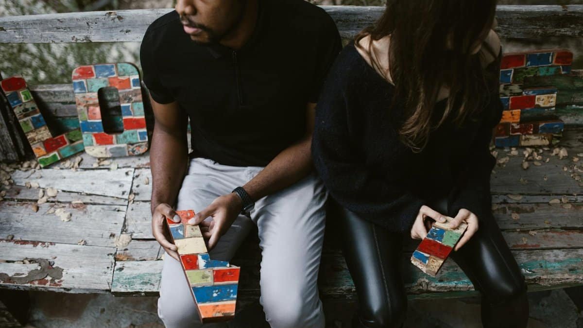 Couple sitting on a bench with colorful blocks forming the word 'LOVE,' embodying complex emotions.