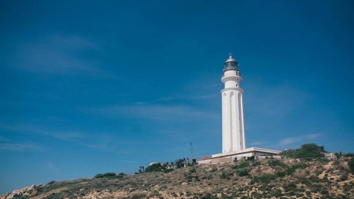 A striking white lighthouse stands tall on a rugged hillside under a vivid blue sky, providing guidance and safety.