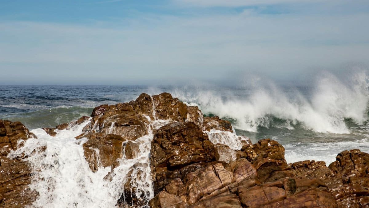 Powerful ocean waves crash against rugged brown rocks under a clear blue sky.