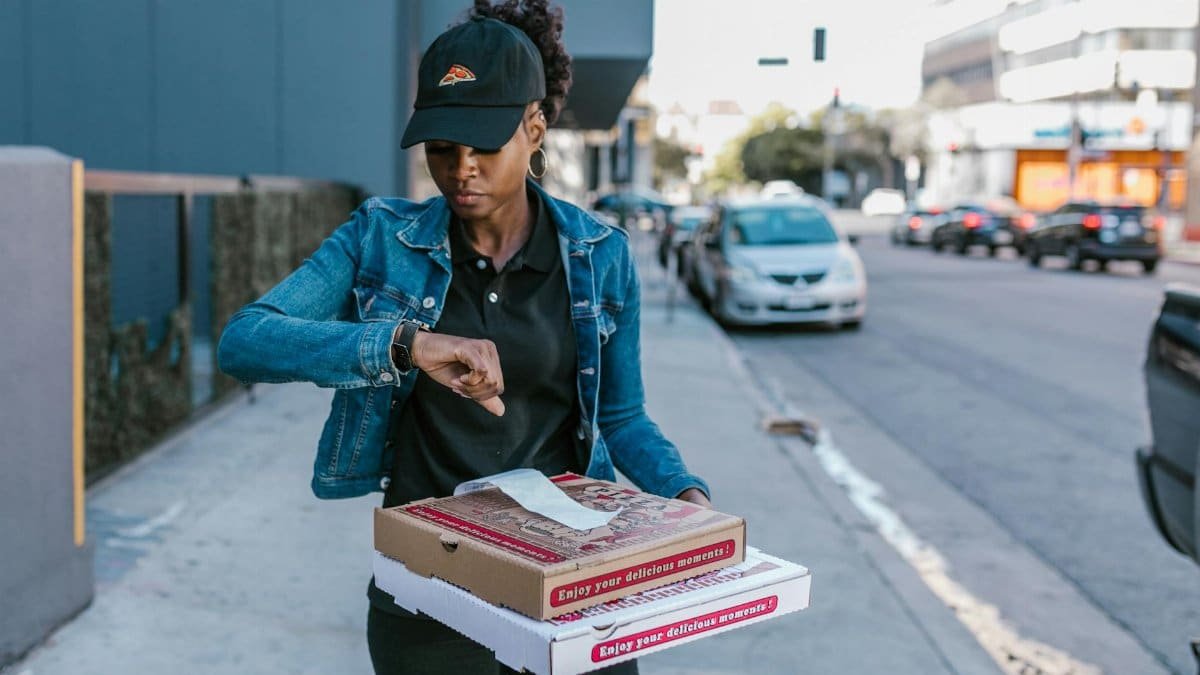 Pizza delivery woman checks time while holding pizza boxes on a city sidewalk.