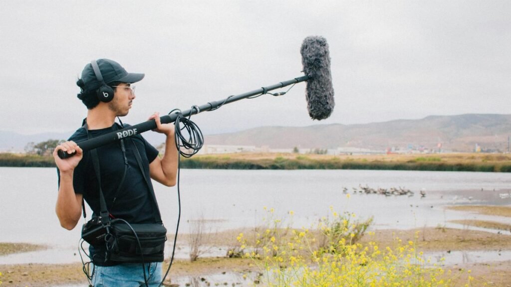 A sound technician operates a boom microphone near a lake with scenic hills in the background.
