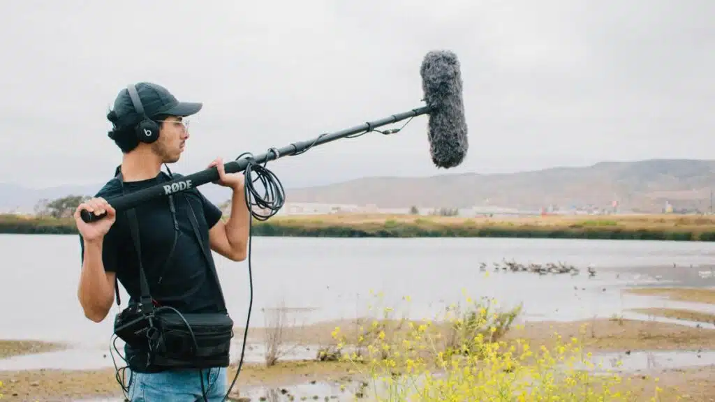 A sound technician operates a boom microphone near a lake with scenic hills in the background.