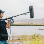 A sound technician operates a boom microphone near a lake with scenic hills in the background.