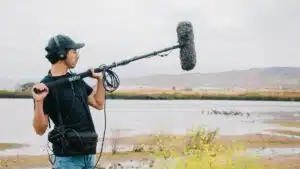 A sound technician operates a boom microphone near a lake with scenic hills in the background.