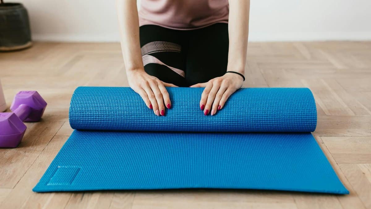 Female preparing for workout by rolling a blue yoga mat on wooden floor with dumbbells nearby.