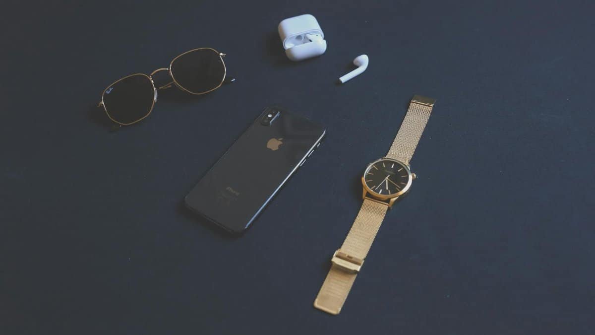 Flat lay of stylish accessories including a smartphone, gold watch, sunglasses, and earphones on a dark background.