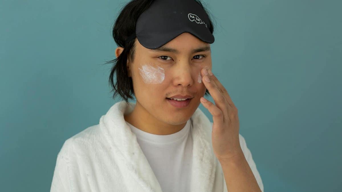 Asian man in a robe applying skincare face cream for facial care with a sleep mask.