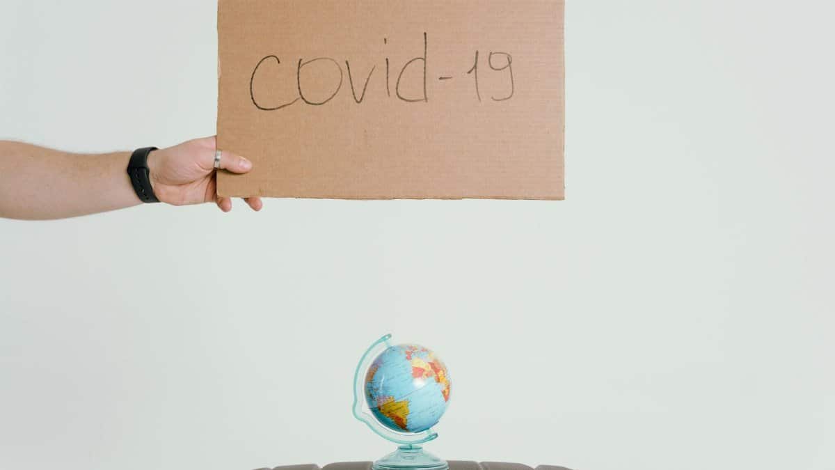 Hand holding covid-19 sign over globe and luggage, symbolizing pandemic travel impact.