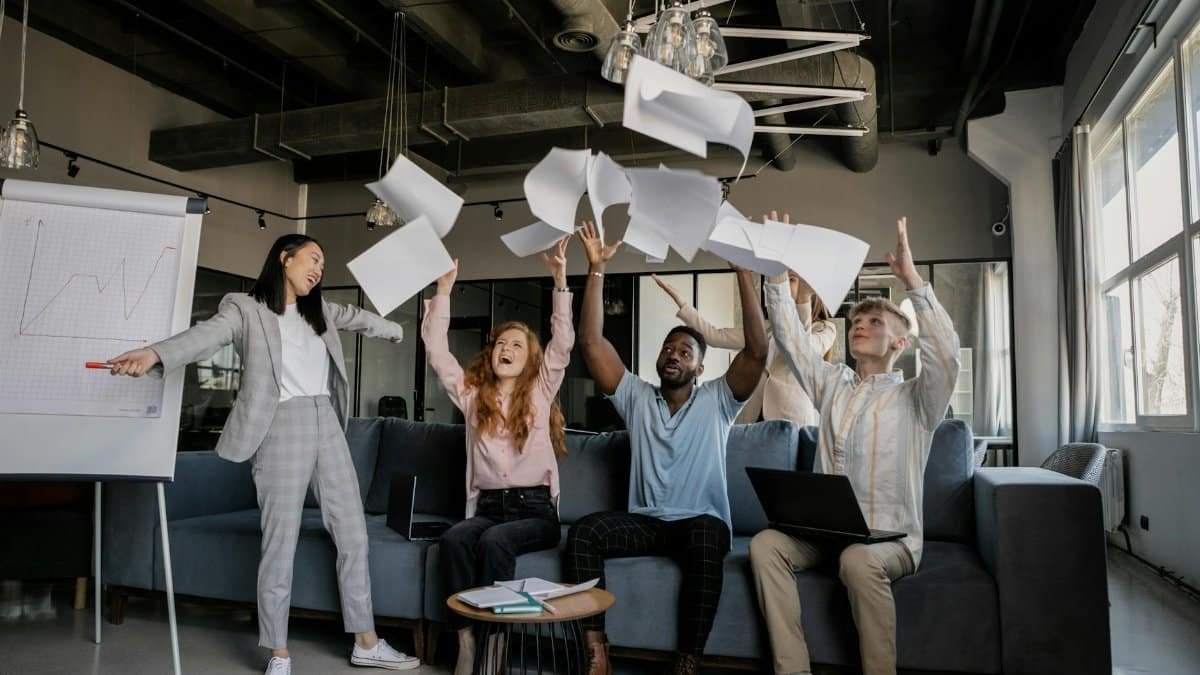Diverse coworkers celebrate success by throwing papers in modern office setting.