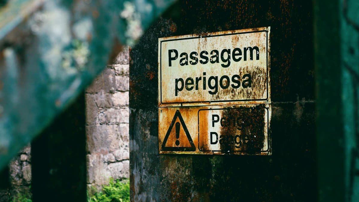 Old rusty warning sign in Portuguese amidst urban decay and vegetation.