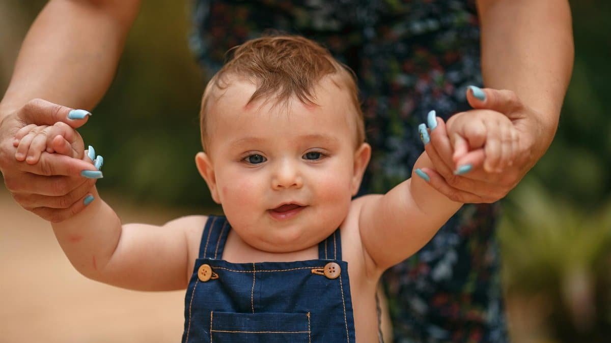 A cute baby guided by an adult as they take their first steps outside on a sunny day.