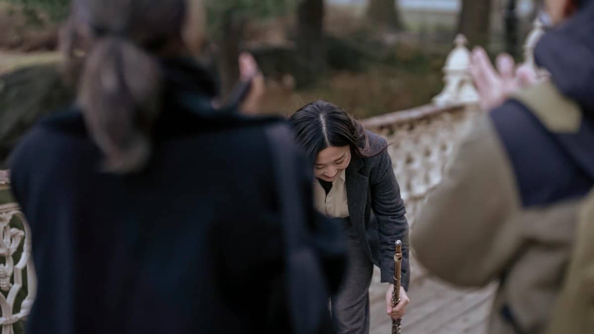 A woman flutist bows on a scenic outdoor path while holding a flute, acknowledging an audience.