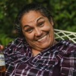 Happy woman smiling broadly while holding a Modelo beer in a garden setting.