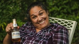 Happy woman smiling broadly while holding a Modelo beer in a garden setting.