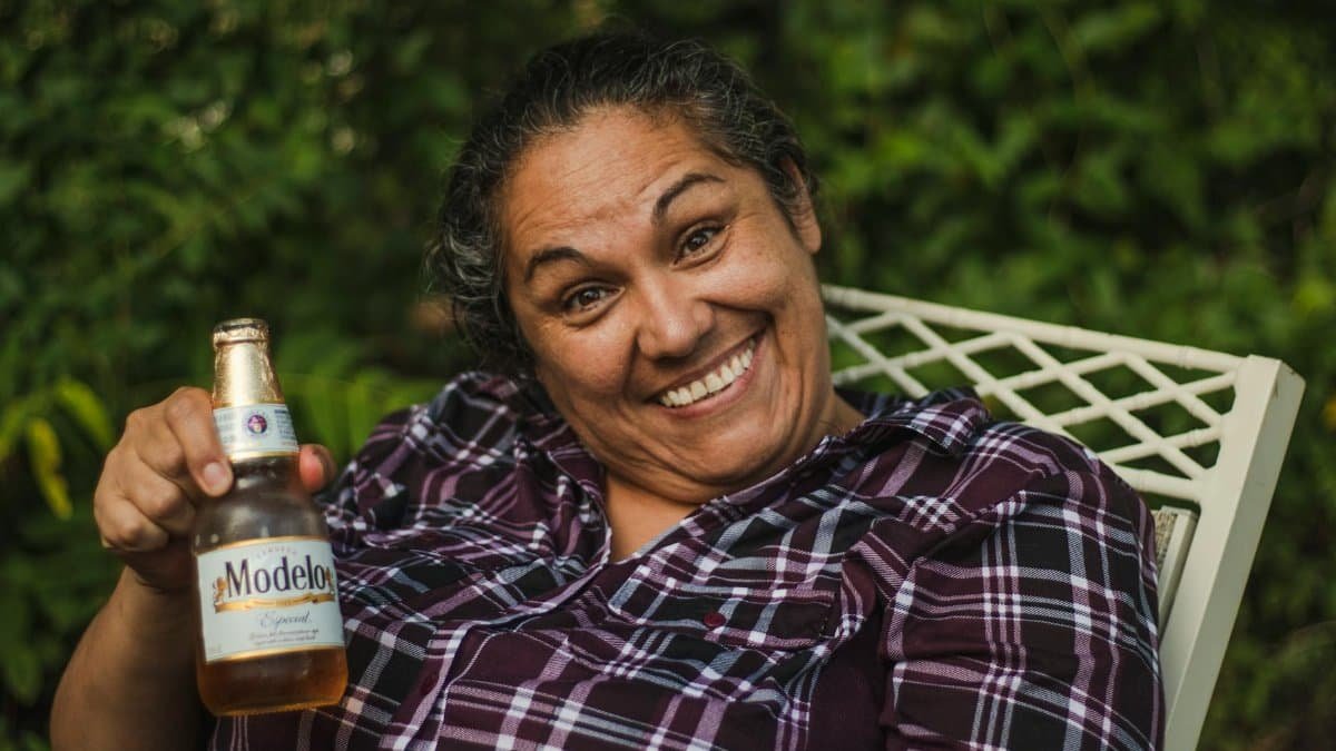 Happy woman smiling broadly while holding a Modelo beer in a garden setting.
