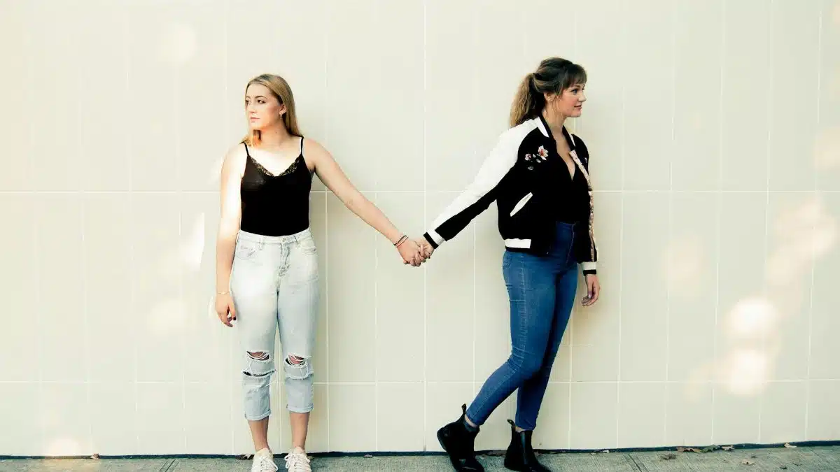 Two young women in casual outfits holding hands against a white tiled wall. Stylish and intimate moment captured.