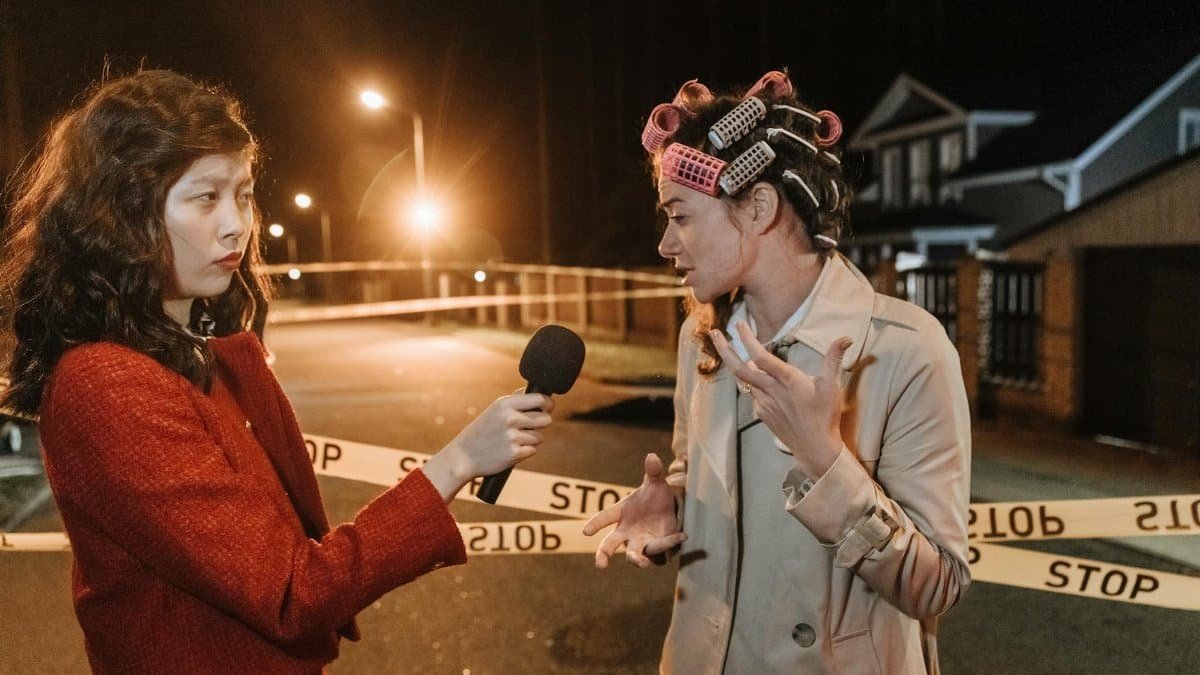 Asian female journalist interviews a woman at a night crime scene. Police tape marks the area.