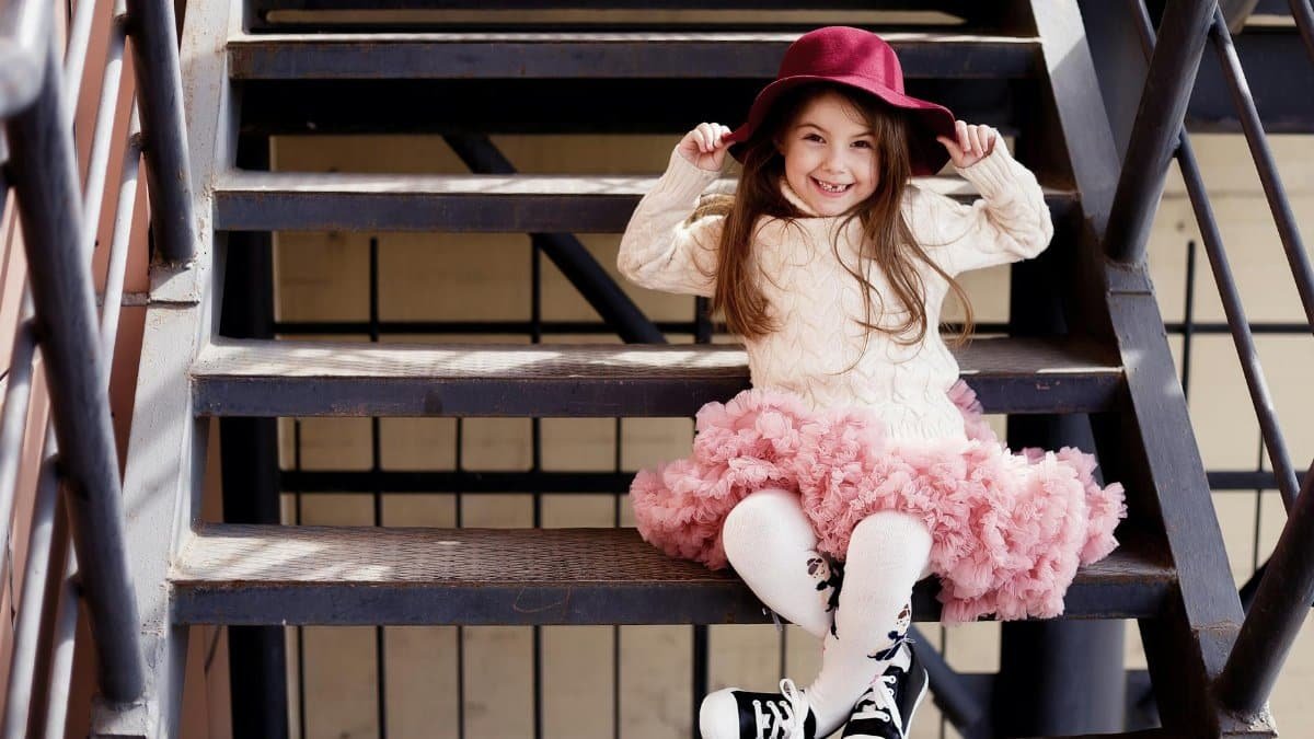 Full body of happy girl in stylish outfit sitting on metal stairway while holding hat and looking at camera