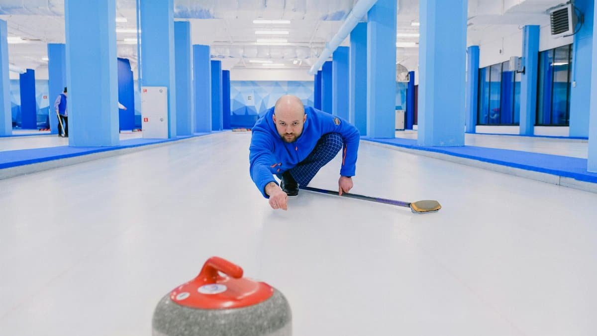 Concentrated sportsman with beard in blue uniform squatting down and looking at sliding stone with red handle during curling game on modern ice rink