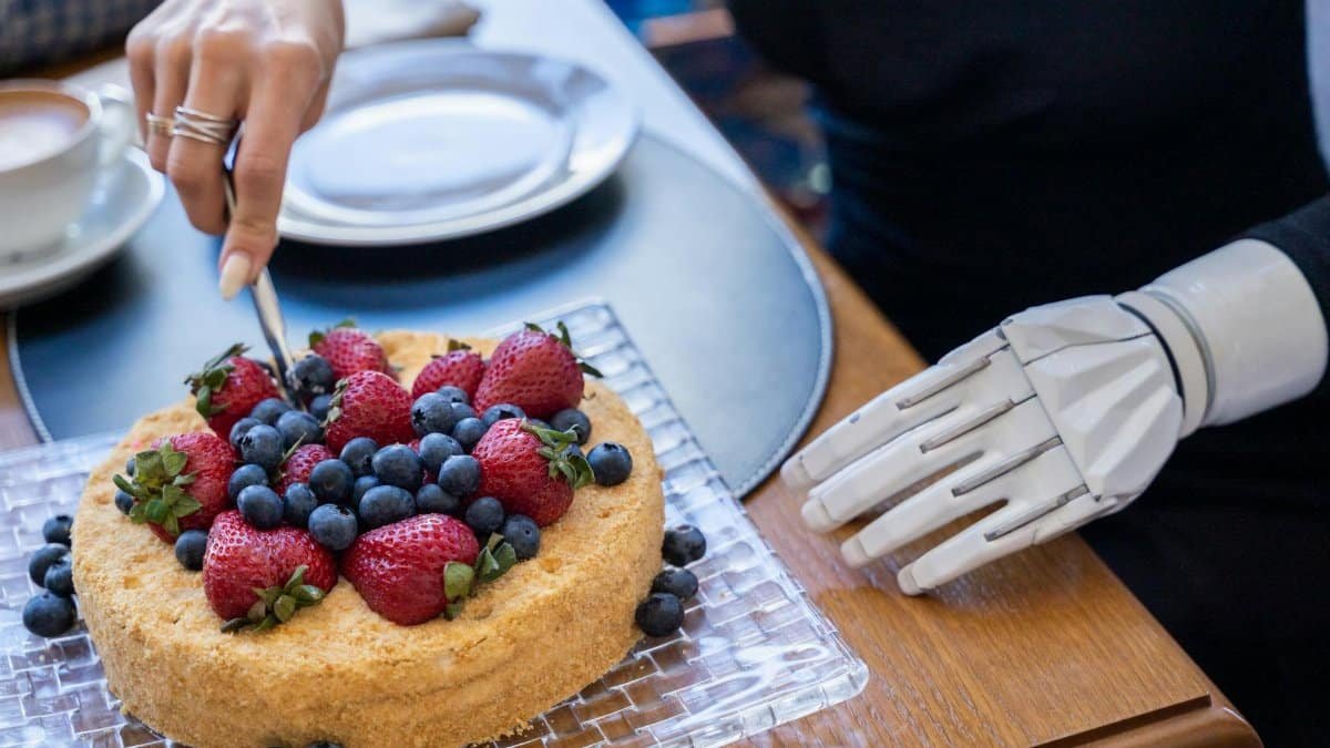 A person with a prosthetic arm cuts a cake topped with strawberries and blueberries, showcasing modern technology.