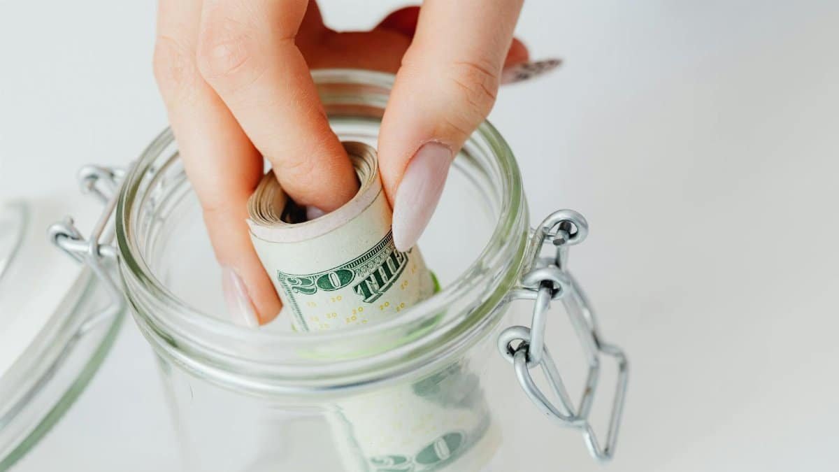 A close-up of a hand placing rolled dollars into a glass jar, symbolizing savings.