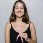 Smiling woman holding a pink ribbon symbolizing breast cancer awareness against a neutral background.