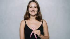 Smiling woman holding a pink ribbon symbolizing breast cancer awareness against a neutral background.