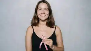 Smiling woman holding a pink ribbon symbolizing breast cancer awareness against a neutral background.
