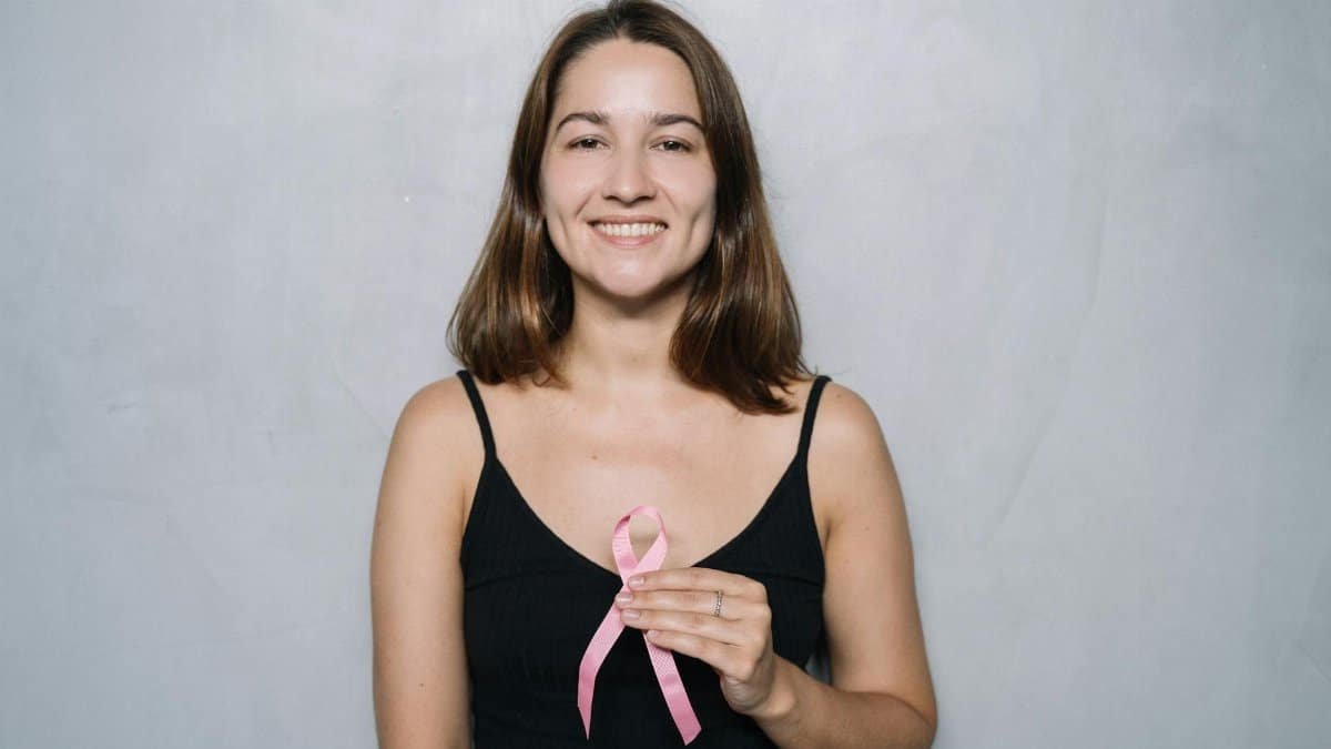 Smiling woman holding a pink ribbon symbolizing breast cancer awareness against a neutral background.