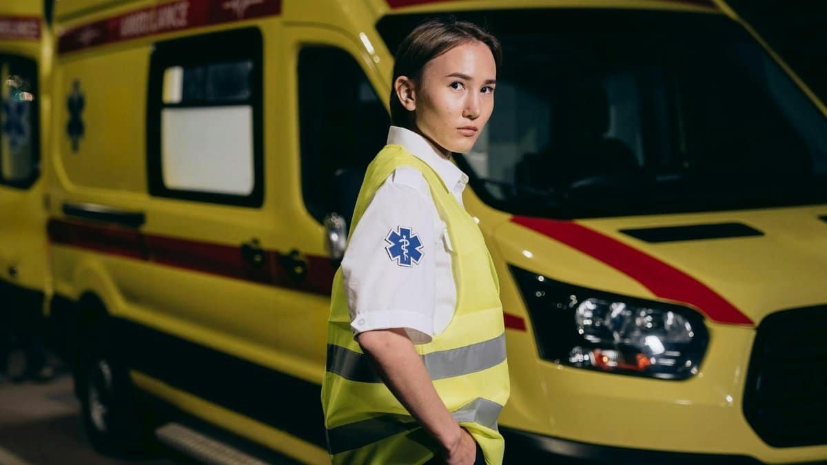 A female paramedic in a reflective vest stands beside a yellow ambulance at night.